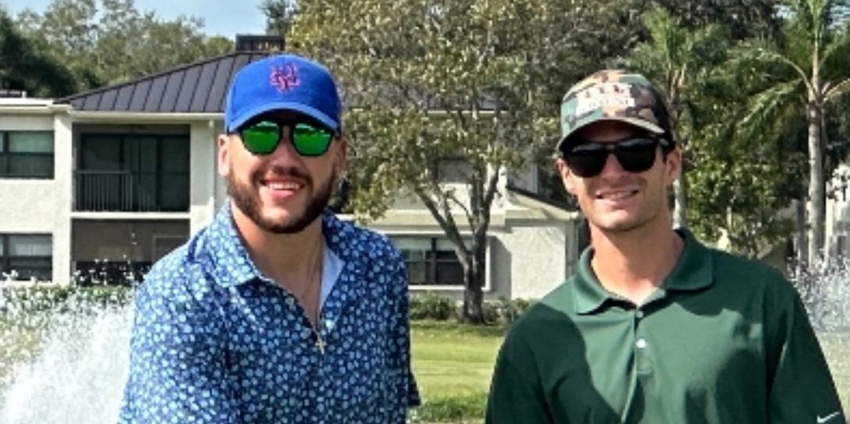 Two men wearing caps and sunglasses smiling outdoors by a water fountain.