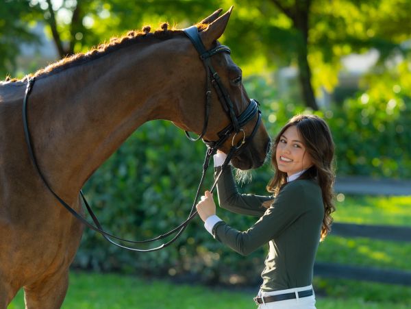 A smiling young woman holding the reins of a brown horse outdoors.