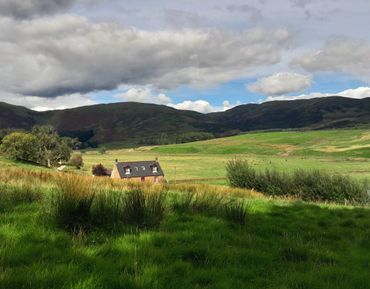 A cozy house nestled in lush green fields under a partly cloudy sky and distant hills.