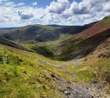 Sunlit green valley surrounded by rolling hills under a partly cloudy sky.