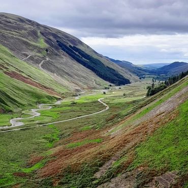 A winding road near moffat through a lush green valley between rolling hills under a cloudy sky.