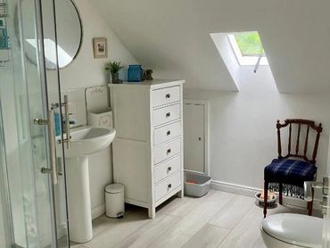 Bright attic bathroom with shower, white furniture, and a wooden chair.