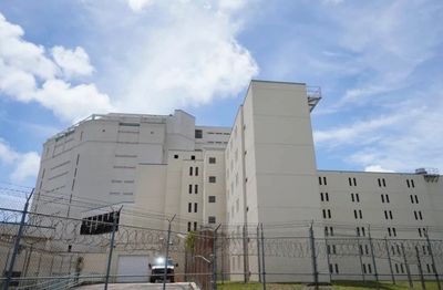 Large beige prison building with barbed wire fence under a cloudy blue sky.