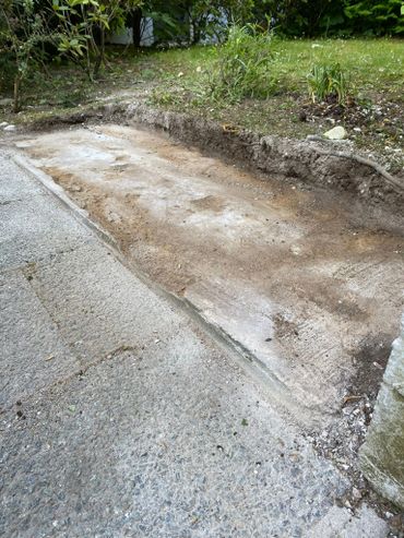 A dug-out rectangular patch of dirt beside a concrete path with some plants in the background.
