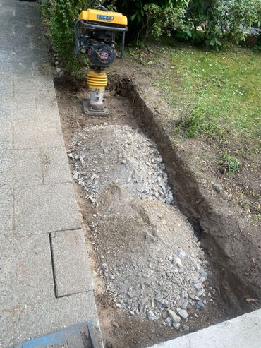 A soil compactor on a dug trench with gravel and soil beside a sidewalk.