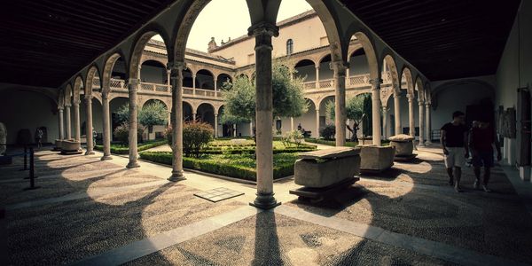 The serene courtyard of the University of Salamanca, with arches, columns, a garden, and visitors.