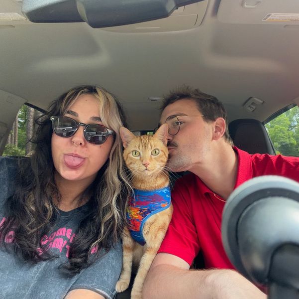 Couple with a cat in a colorful harness inside a car, sharing a playful moment.