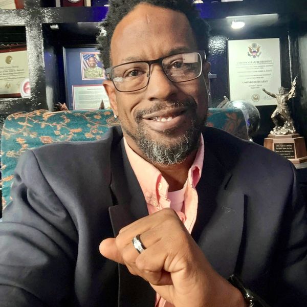 Smiling man wearing glasses and a suit jacket, showing a ring on his hand.