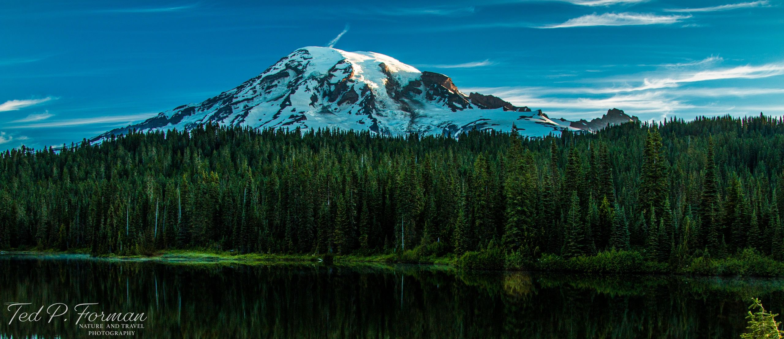 Mount Rainier peeking behind pine tree forest and reflected in a lake