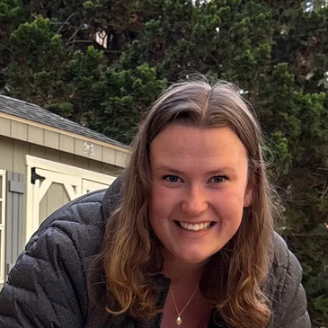 Smiling woman outdoors in casual jacket in front of a shed and trees.