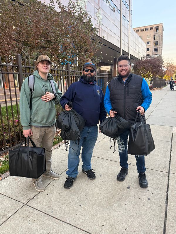 Three men standing on a sidewalk holding bags, smiling at the camera.
