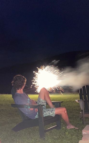 Person sitting on a chair with fireworks sparking behind them at night.