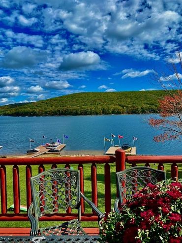Scenic lake view from a wooden deck with chairs and flowers under a partly cloudy sky.