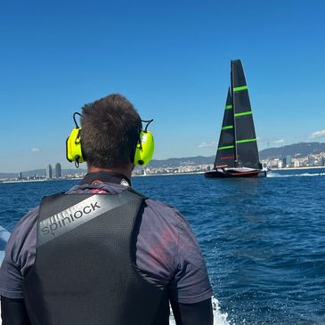 Man with yellow earmuffs watches a sleek black and green sailboat on the sea.