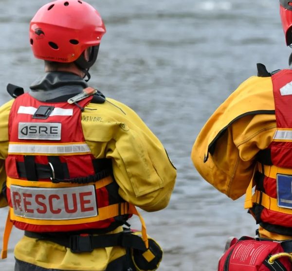 Two rescue workers in yellow suits and red helmets near water.