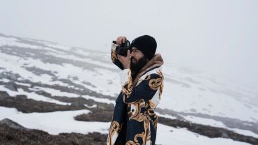 Man in patterned coat taking a photo in snowy landscape.