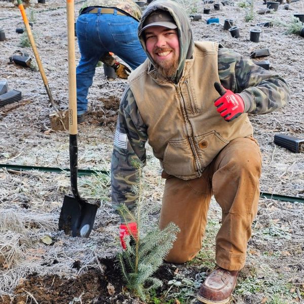 Man planting a small tree, smiling and giving thumbs up outdoors.