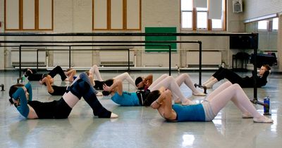 Dancers in class laying on the floor with knees bent and heads resting in their hands.