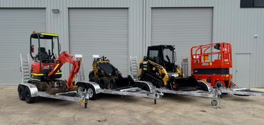 Four different types of construction equipment on trailers outside a warehouse.