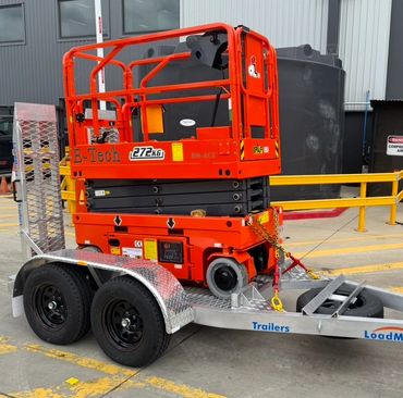 Orange scissor lift secured on a dual-axle trailer in an industrial area.