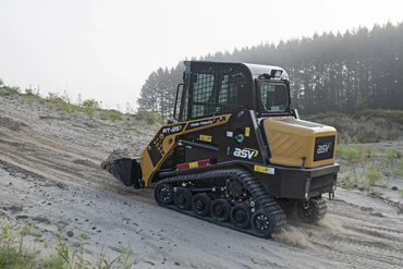 Tracked skid-steer loader moving uphill on a sandy terrain.