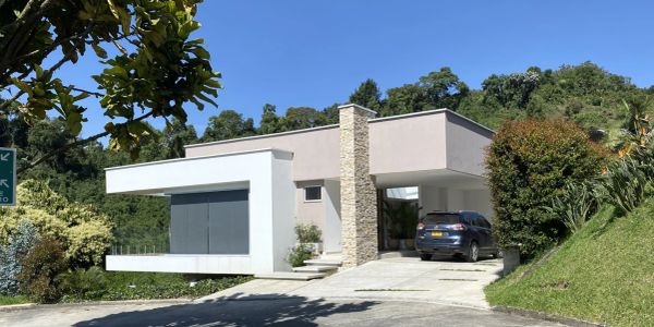 Modern house with stone chimney and carport in lush green surroundings.