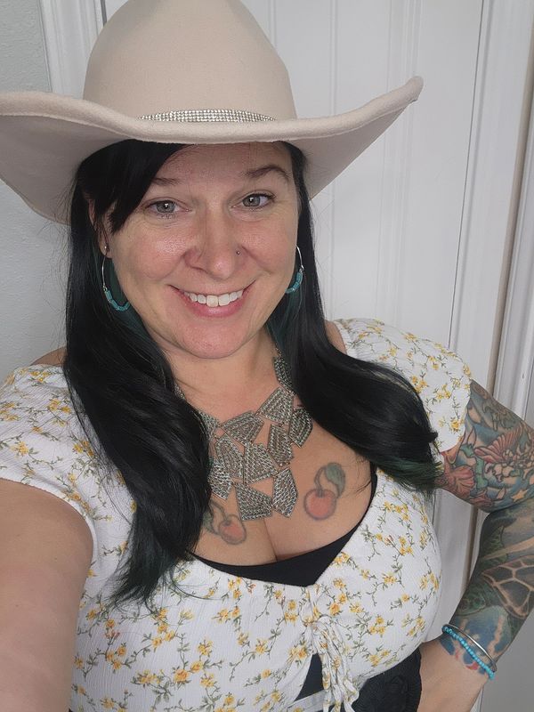 Smiling woman in a cowboy hat with tattoos and floral top.