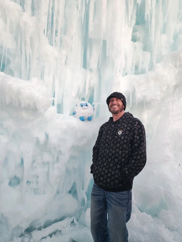 Man in a black hoodie smiles inside an ice cave with icicles and a small yeti toy.