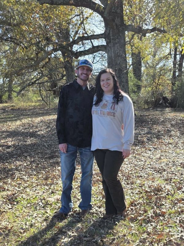 A couple smiling outdoors in a wooded area during autumn.