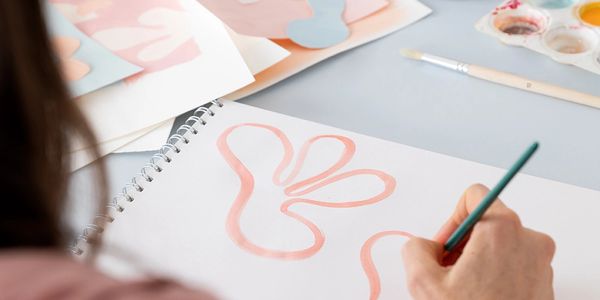 Person painting an abstract pink floral on paper, with brushes and cut-out shapes scattered nearby.