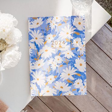 2024 teacher planner with blue daisy design on outdoor table beside vase of white flowers.
