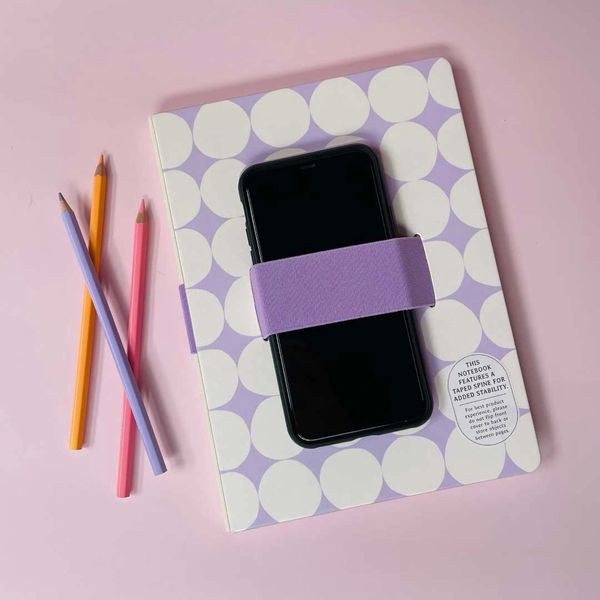 Lavender polka dot notebook with elastic band phone holder beside pastel pencils on pink background.