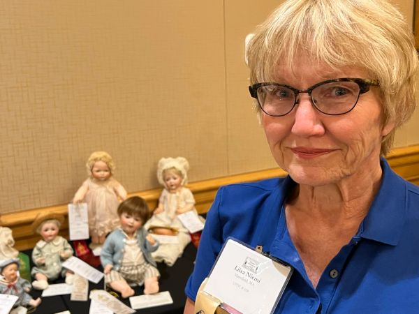 Woman with glasses posing with collectible dolls in the background.