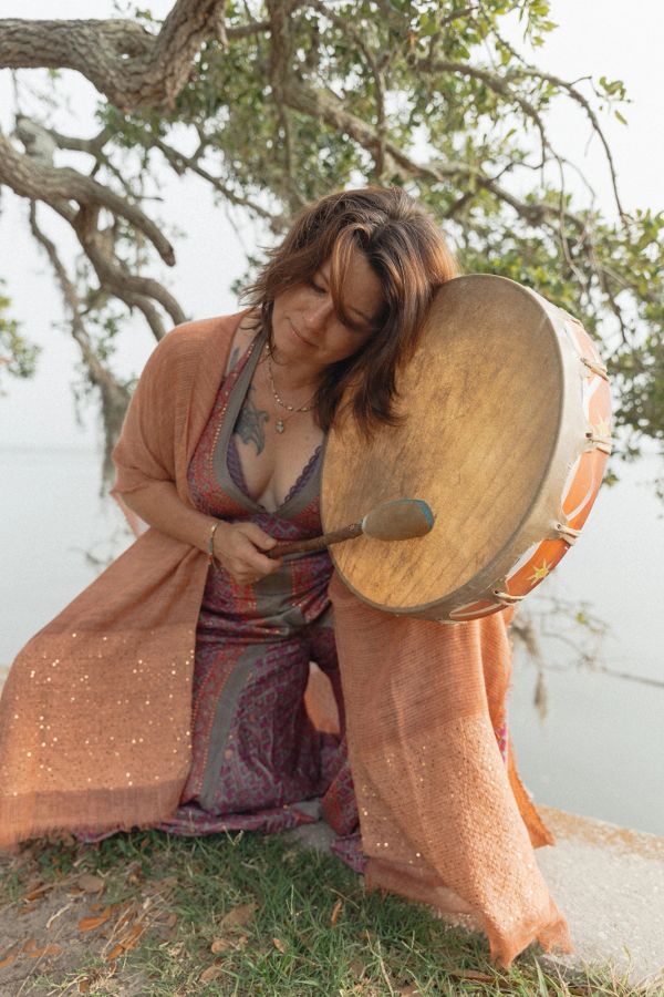 Woman playing a large drum outdoors near a tree.