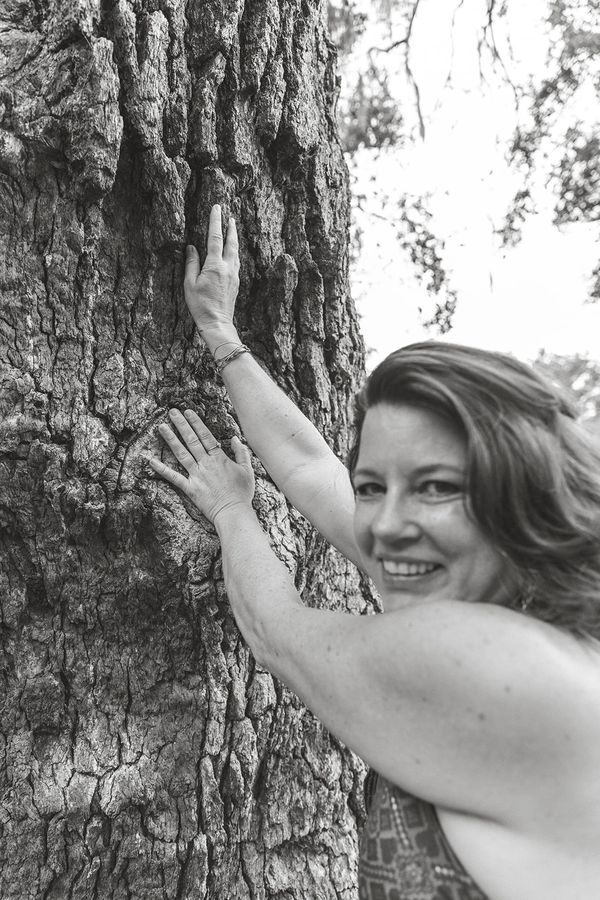 Woman smiling while embracing a large tree trunk outdoors.