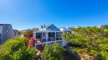 White two-story house with a balcony surrounded by lush greenery and flowers under a clear blue sky.