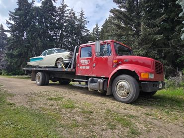 Red tow truck carrying a classic car on a forest road.