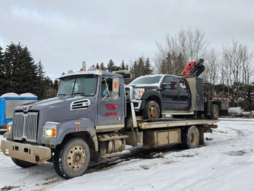 Tow truck carrying a damaged black pickup truck in a snowy area.
