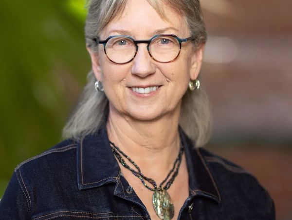 Smiling woman with glasses and necklace in denim jacket.