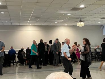 Seniors socializing and dancing in a community hall with tables and chairs.
