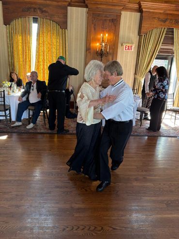 Elderly couple dancing closely in an elegant room with seated guests.