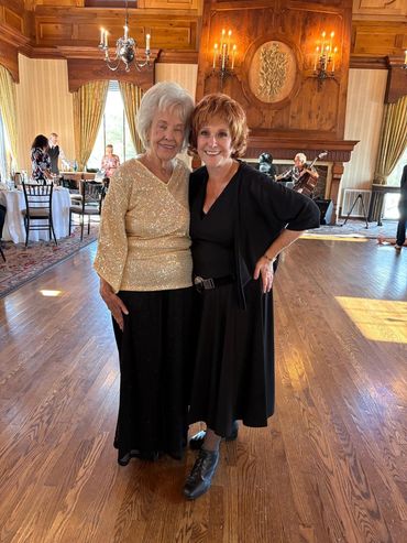 Two elderly women smiling and posing in a warmly lit, elegant room.