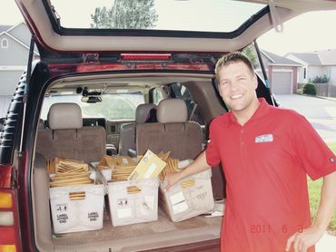 Man in red shirt smiling beside an SUV trunk filled with mail bins.