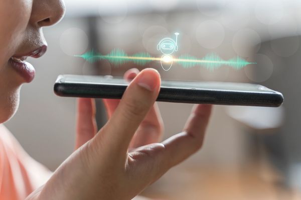 A person speaks into a smartphone with a chatbot icon and sound waves above it.