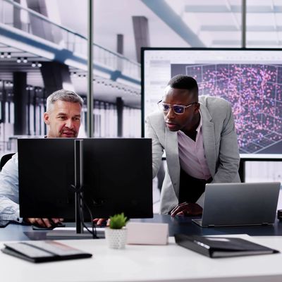 Two colleagues working on computers in a modern office, with a large screen displaying data.