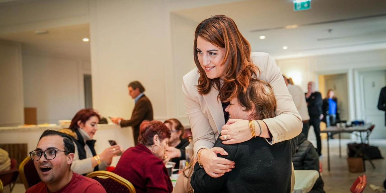 A woman in a beige blazer warmly hugs another person in a lively indoor gathering.