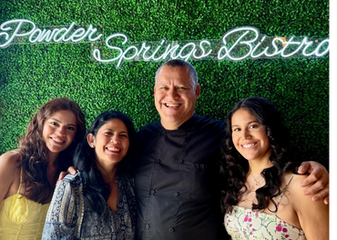 Four smiling people posing in front of a green wall with a neon sign.