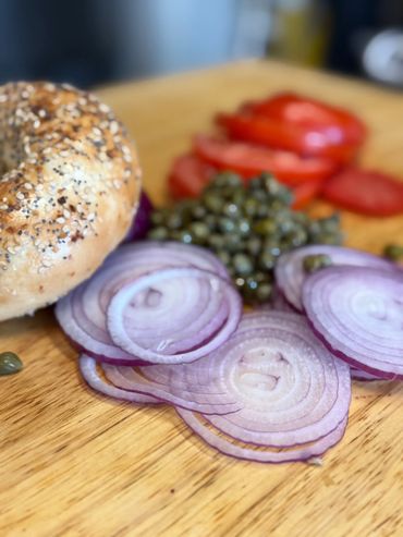 Sliced red onions, capers, tomato slices, and an everything bagel on a wooden board.