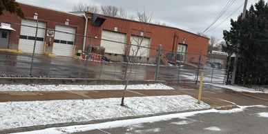 A snowy sidewalk and street in front of a fenced industrial building with garage doors.