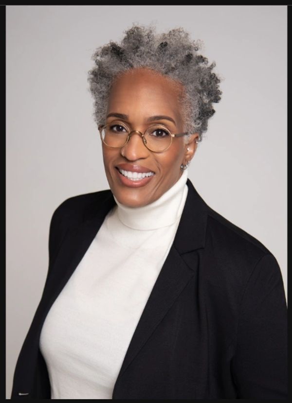 Smiling woman with gray curly hair, glasses, white turtleneck, and black blazer.